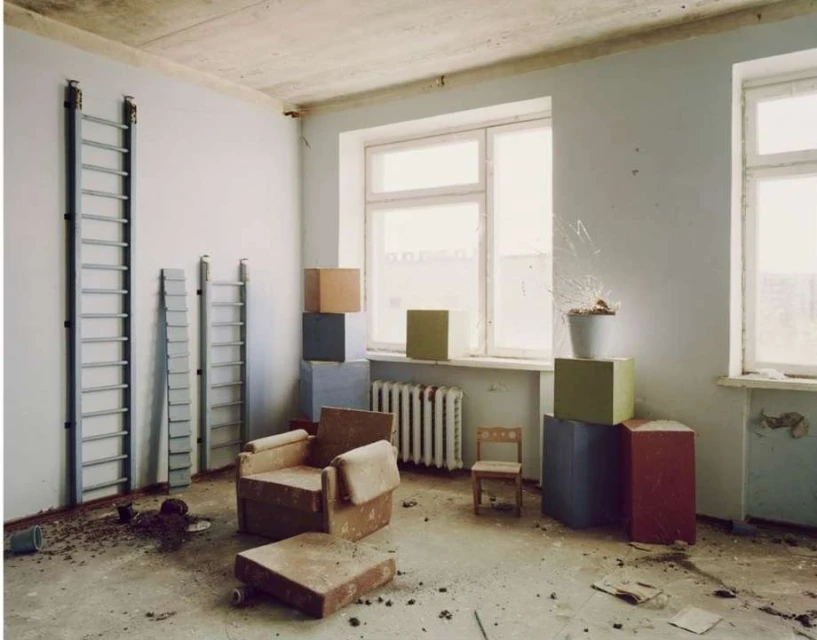 A vacant indoor room features a worn armchair, radiator, ladders on the wall, and potted plants, bathed in soft, diffused natural light.