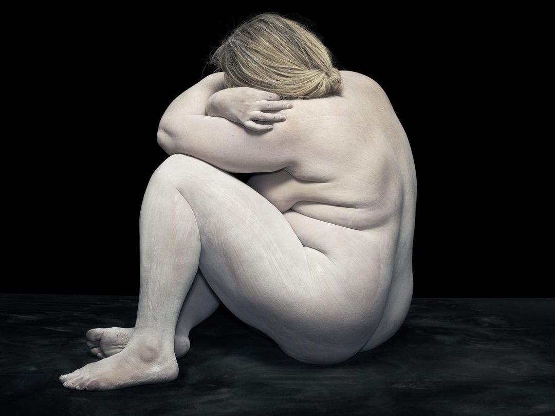 Nude adult female with painted skin sits hunched with arms over head, posing against a black background.