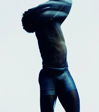 Profile of a black man with curly hair posing with arms raised overhead against a bright white background