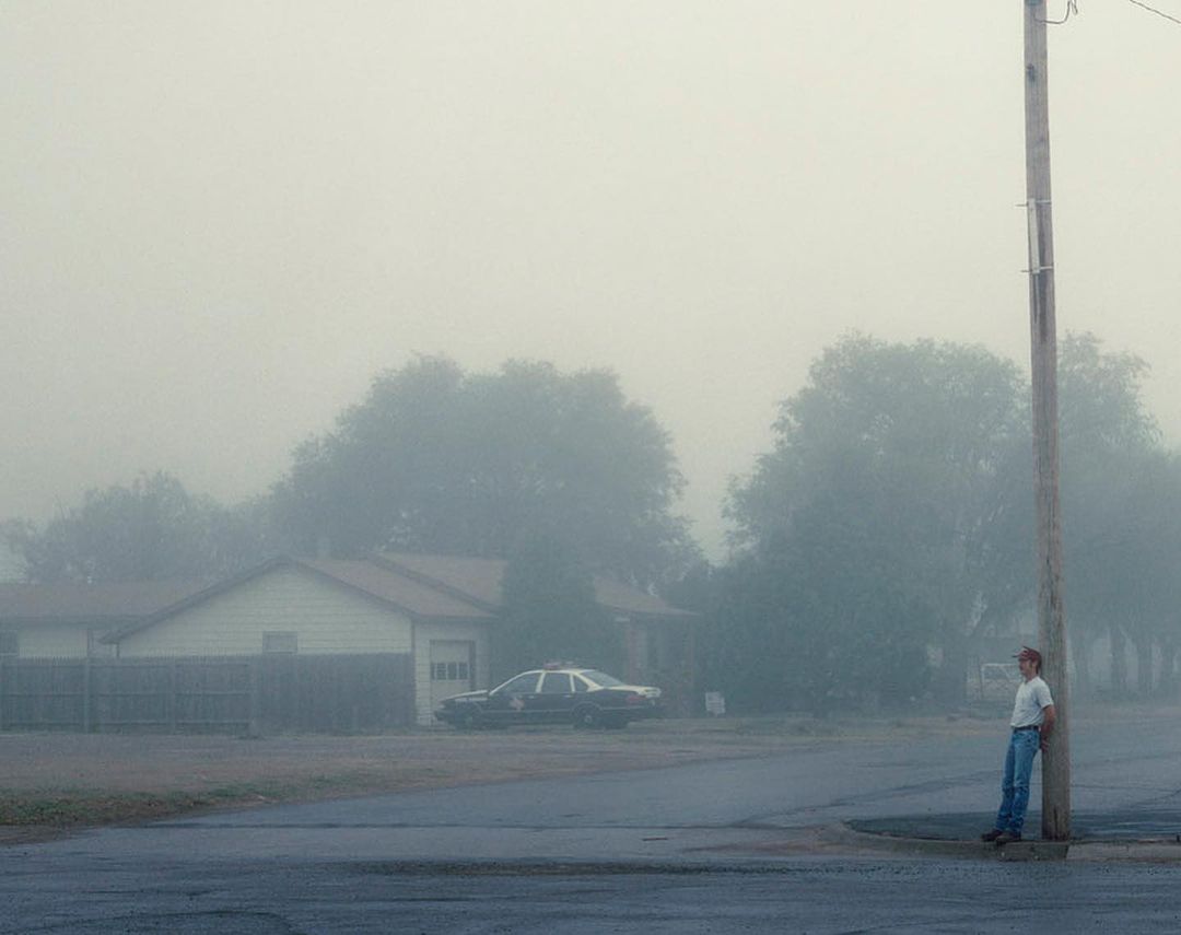 A young adult leans against a pole on a foggy street near a parked police car, creating a somber, street scene with diffused light.