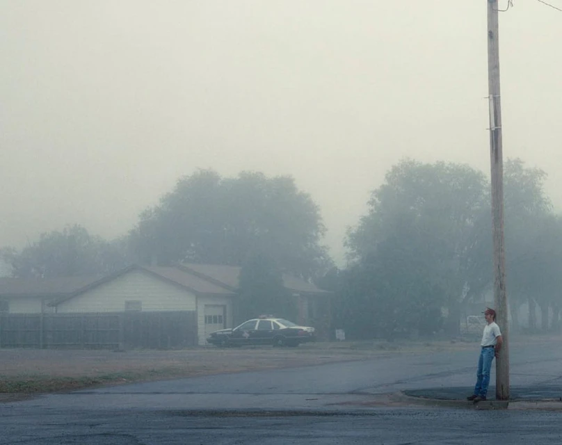 A young adult leans against a pole on a foggy street near a parked police car, creating a somber, street scene with diffused light.
