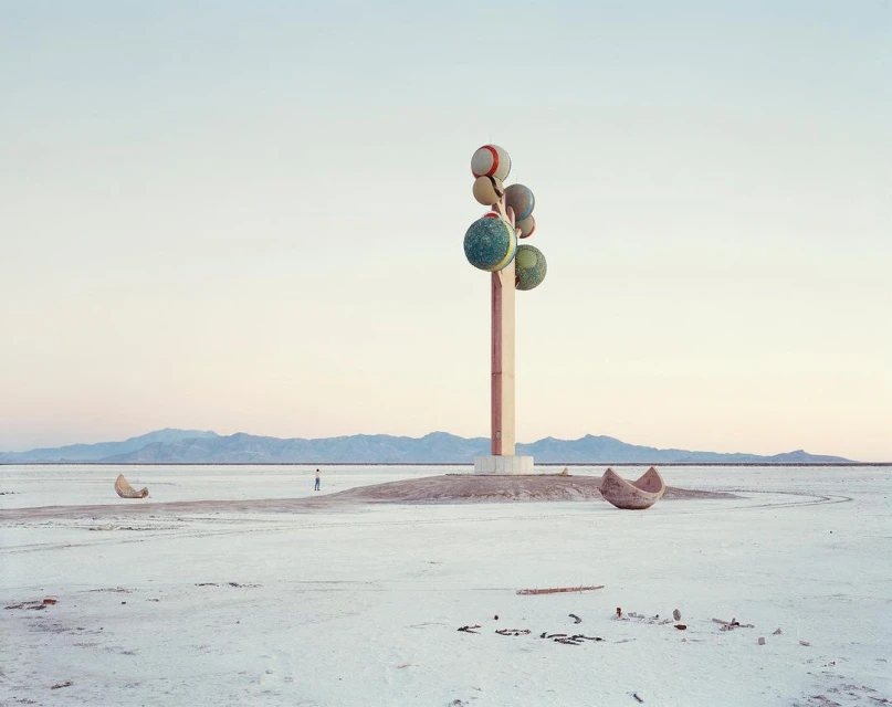 Outdoor scene with a person in the distance, a tall sculpture with spheres, and two boat-shaped sculptures on a flat, desert-like landscape.