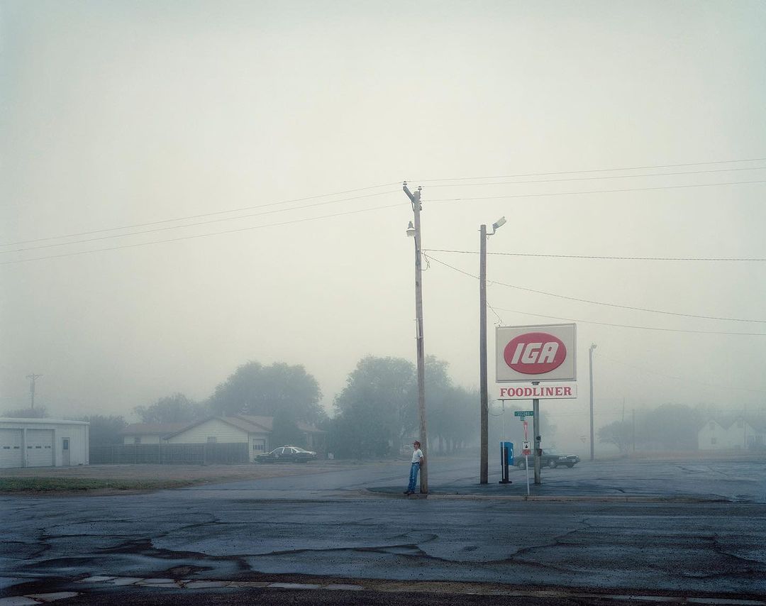 Foggy street scene featuring a lone person standing near an IGA grocery store sign, with muted tones and a quiet, rural atmosphere.