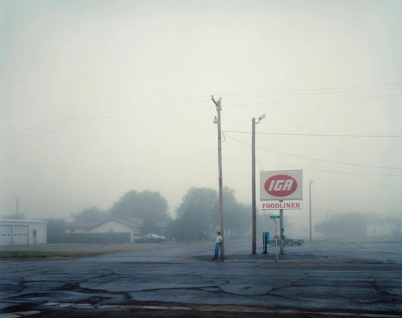 Foggy street scene featuring a lone person standing near an IGA grocery store sign, with muted tones and a quiet, rural atmosphere.