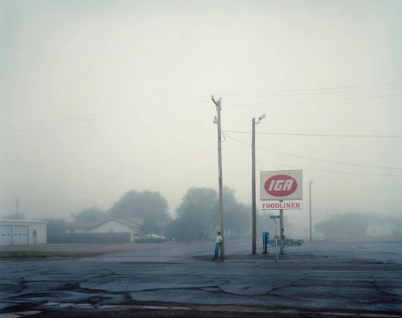 Foggy street scene featuring a lone person standing near an IGA grocery store sign, with muted tones and a quiet, rural atmosphere.