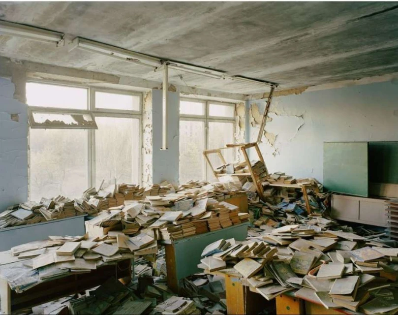 Interior view of abandoned classroom with scattered books, desks, damaged walls, and bright windows suggesting post-apocalyptic scene.
