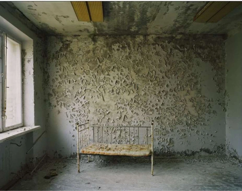 Abandoned room with peeling paint and a metal bed frame. Window light illuminates the decay in this evocative interior photograph.