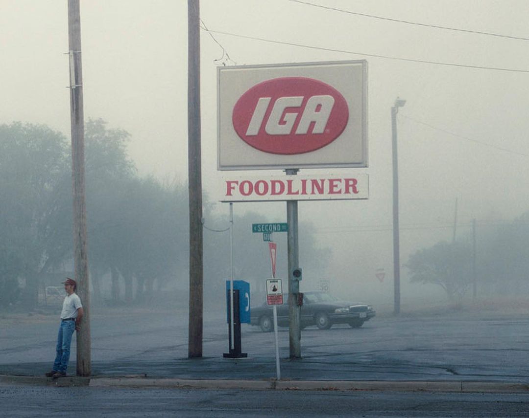 Man leans by sign of IGA Foodliner on a foggy day. Classic car is parked behind him, trees are in background with dense fog.