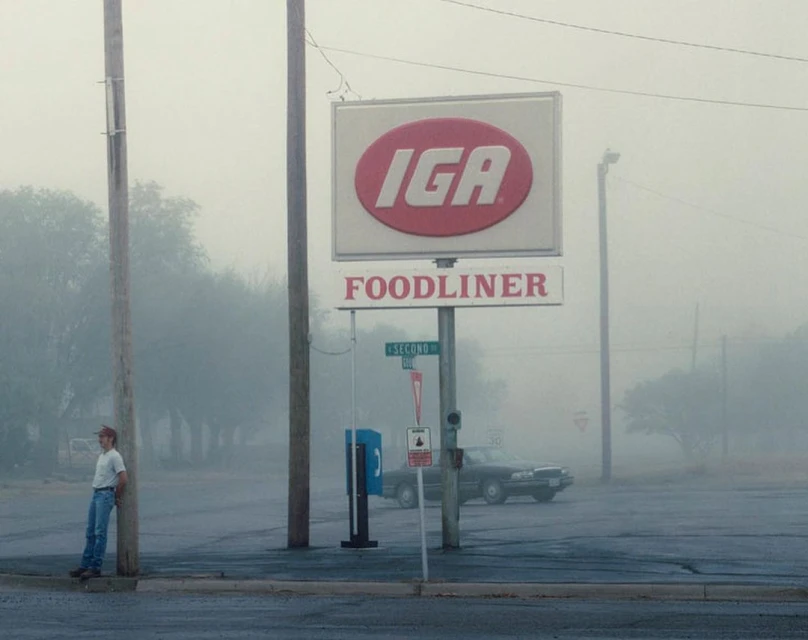 Man leans by sign of IGA Foodliner on a foggy day. Classic car is parked behind him, trees are in background with dense fog.