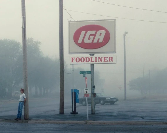 Man leans by sign of IGA Foodliner on a foggy day. Classic car is parked behind him, trees are in background with dense fog.