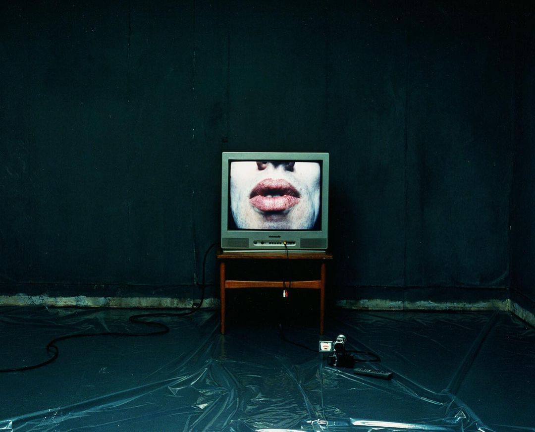 Vintage TV displaying a face against a dark wall on a wood table, with retro camera and black cord on a plastic-covered floor.
