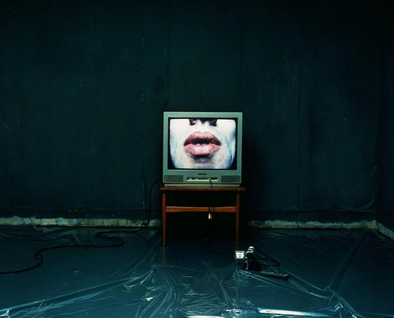 Vintage TV displaying a face against a dark wall on a wood table, with retro camera and black cord on a plastic-covered floor.