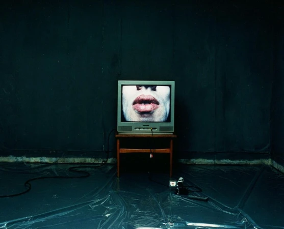 Vintage TV displaying a face against a dark wall on a wood table, with retro camera and black cord on a plastic-covered floor.
