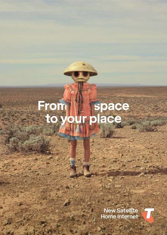 Child stands in a desert landscape wearing an unusual, round-faced mask and a vintage dress in a conceptual advertising image.