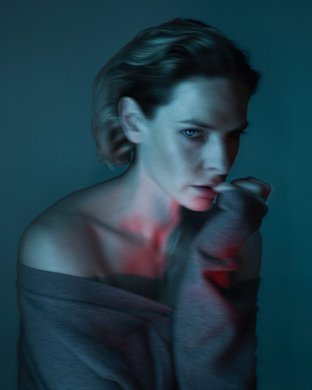 Portrait of a young fair-skinned woman with light brown hair wearing a gray sweater, in cool, soft lighting with a red light accent.