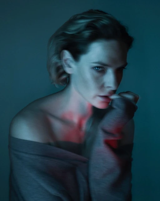 Portrait of a young fair-skinned woman with light brown hair wearing a gray sweater, in cool, soft lighting with a red light accent.