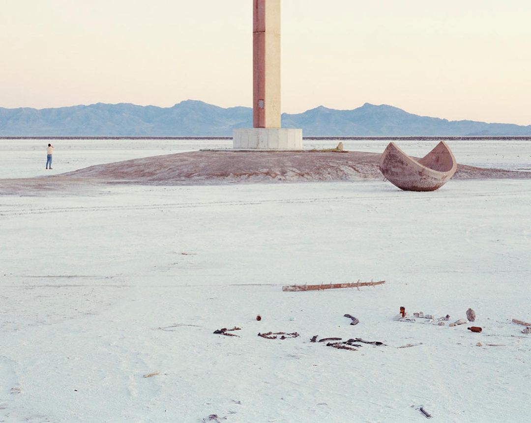 Minimal landscape featuring a lone man near a concrete structure, an upturned boat, and scattered debris on a flat, barren expanse.