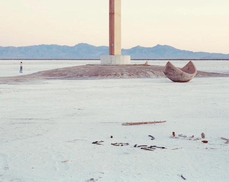 Minimal landscape featuring a lone man near a concrete structure, an upturned boat, and scattered debris on a flat, barren expanse.