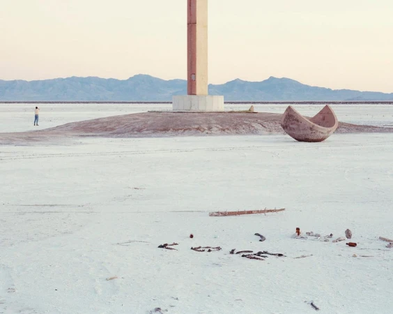Minimal landscape featuring a lone man near a concrete structure, an upturned boat, and scattered debris on a flat, barren expanse.