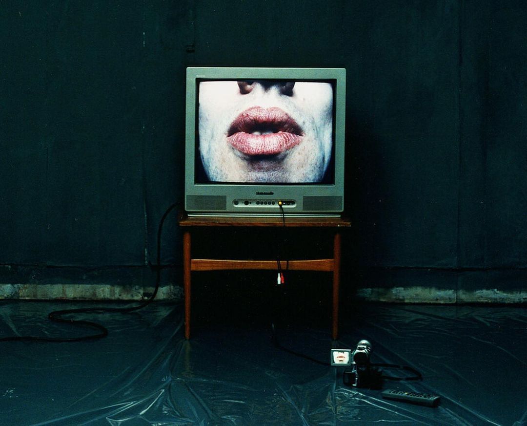 Television displays large lips on a wooden table, situated on a shiny plastic surface with a camcorder and remote nearby. Dark atmosphere.