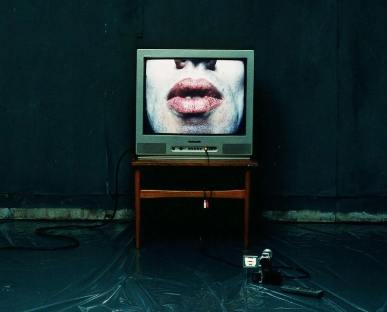 Television displays large lips on a wooden table, situated on a shiny plastic surface with a camcorder and remote nearby. Dark atmosphere.