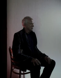 Elderly man in a dark blazer seated on a wooden chair against a light textured wall with subtle red accent lighting