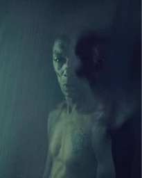 Portrait of a shirtless man with a tattoo on his chest against a textured dark background by photographer Nadav Kander.