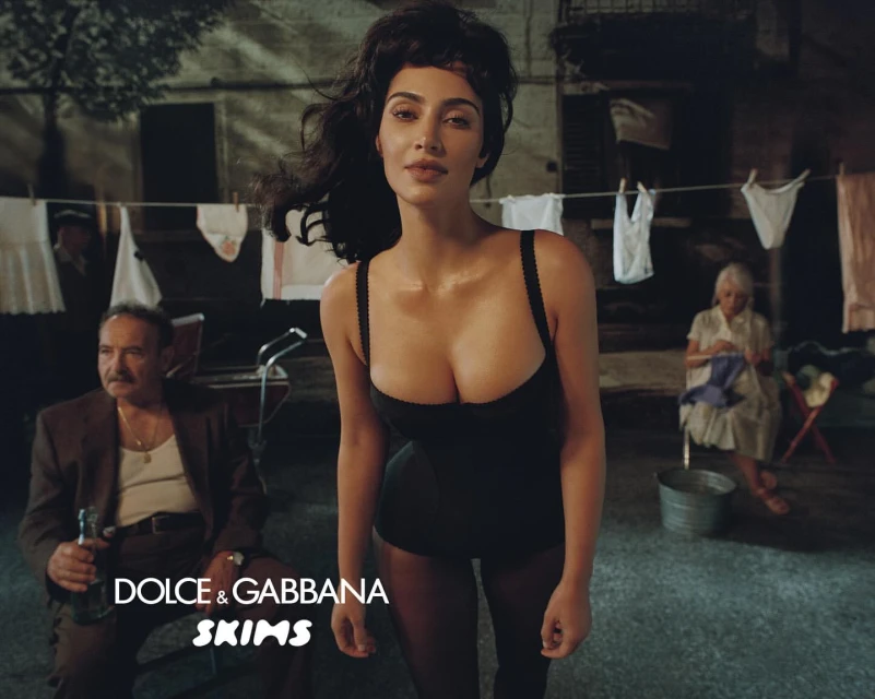 Woman poses in lingerie in a gritty outdoor setting with two older people seated in the background, clotheslines visible, and brand logos.