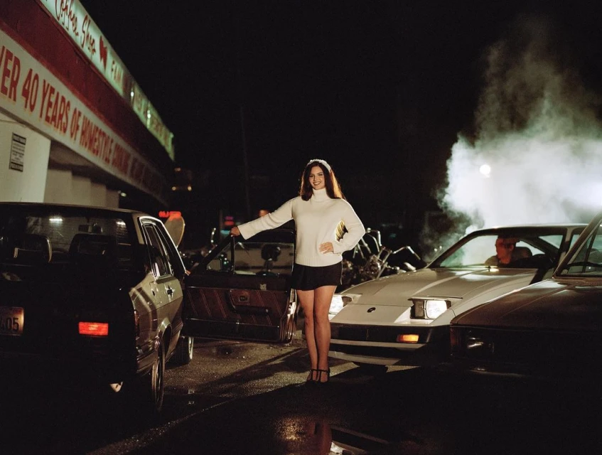 Attractive woman in a white turtleneck sweater and short skirt stands next to cars at night, with a diner visible in the background.