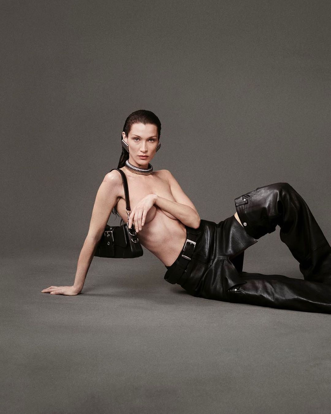 Adult female model poses while reclining in low-rise, loose-fitting black leather pants and purse against grey background in studio shot.