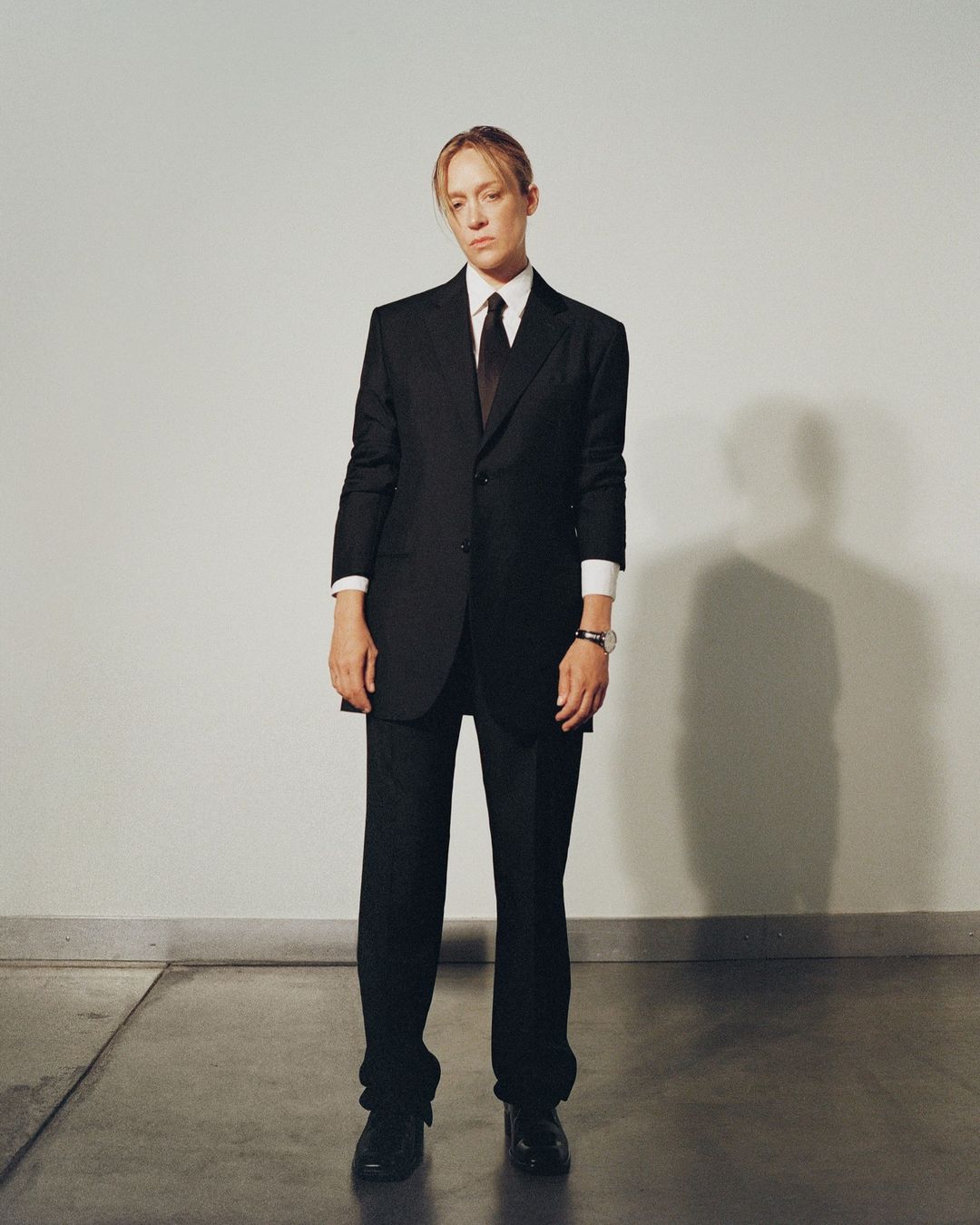 Adult female with neutral expression in a dark business suit with tie standing against a grey wall indoors with polished concrete floors.