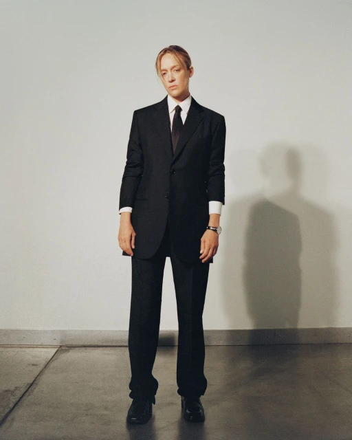 Adult female with neutral expression in a dark business suit with tie standing against a grey wall indoors with polished concrete floors.
