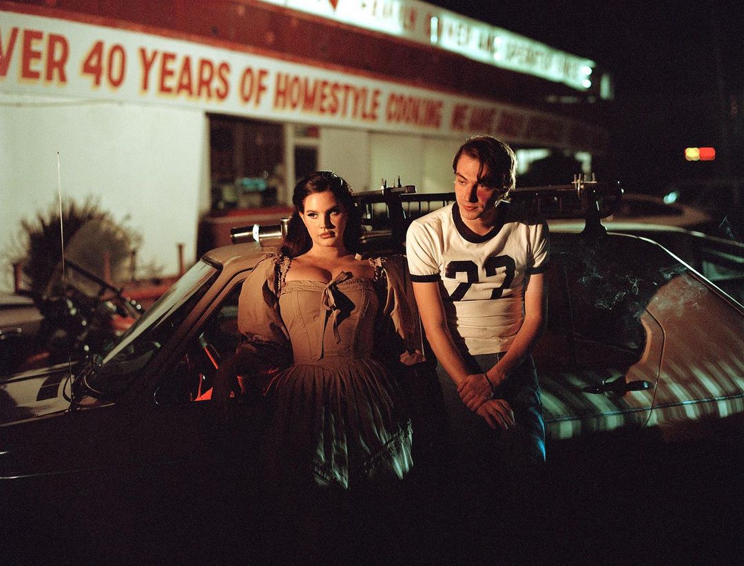 Two people, a man and woman, stand in front of a car. The woman is wearing an old-fashioned dress and the man is wearing a t-shirt.