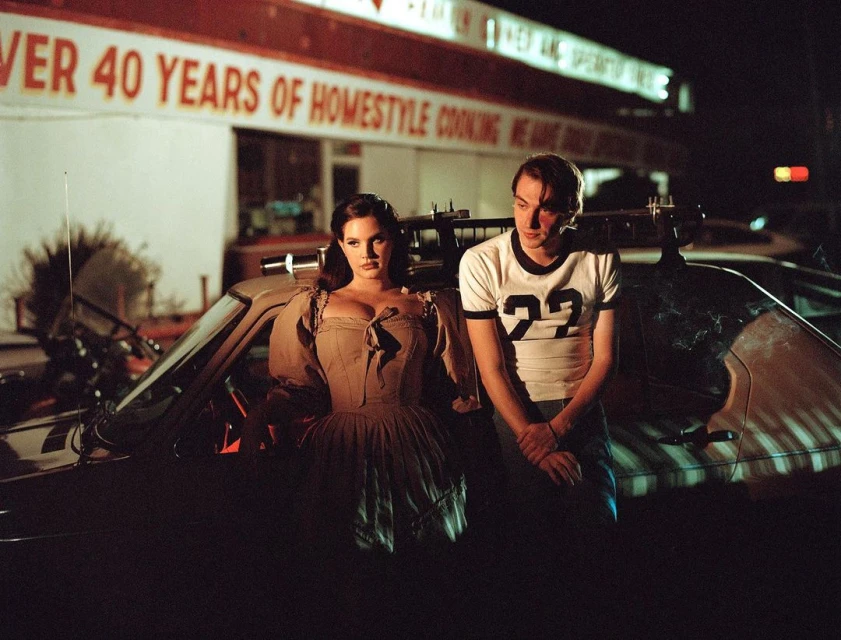 Two people, a man and woman, stand in front of a car. The woman is wearing an old-fashioned dress and the man is wearing a t-shirt.