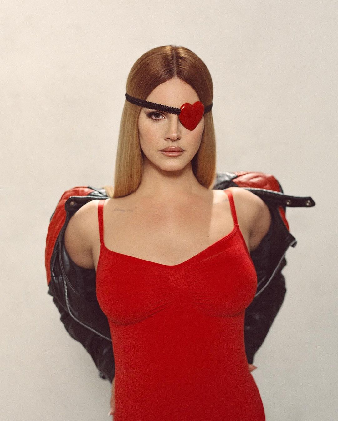Portrait of a blonde woman wearing a red dress and heart-shaped eyepatch, with a black jacket draped over her shoulders against a plain backdrop.