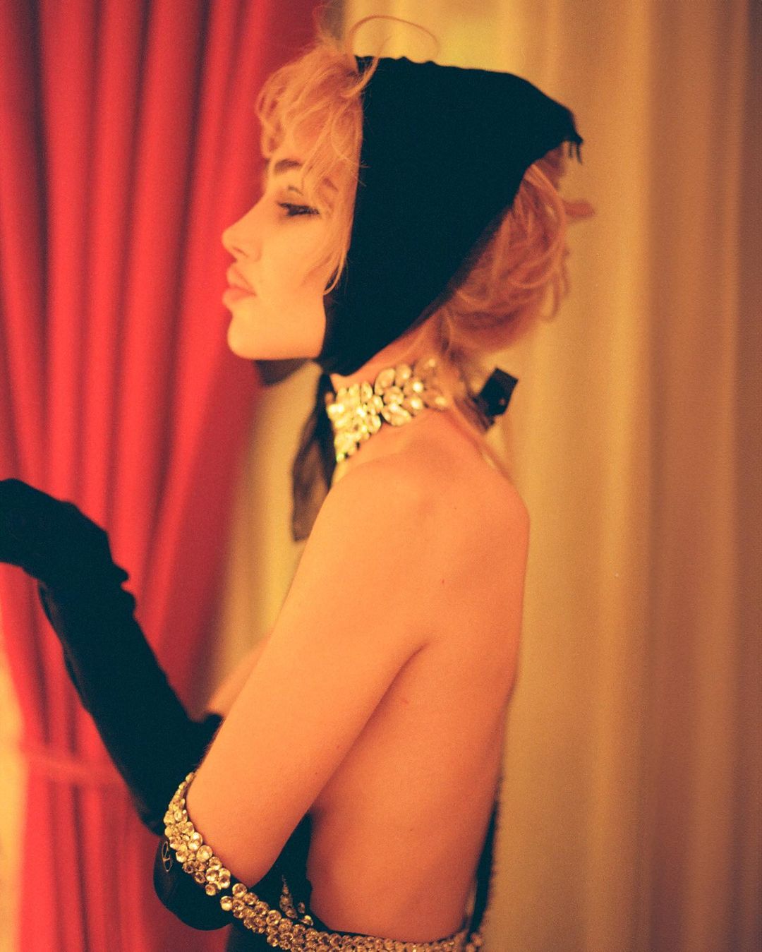 Woman in black headscarf, choker, and gloves poses against red and yellow curtains in a side view, emphasizing vintage glamour.
