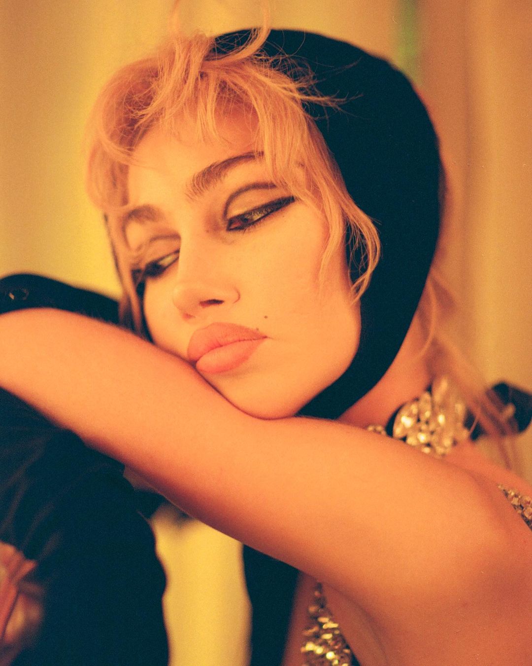 Close-up portrait of a young adult woman with blonde hair, wearing a black headscarf and jewelry, with a warm yellow-orange color grading.