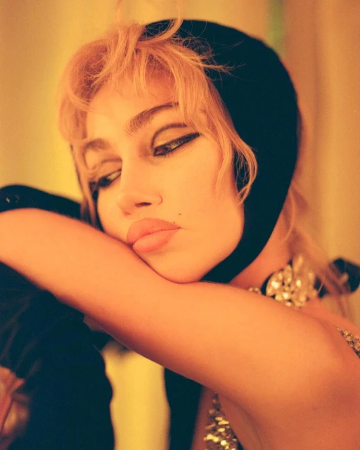 Close-up portrait of a young adult woman with blonde hair, wearing a black headscarf and jewelry, with a warm yellow-orange color grading.