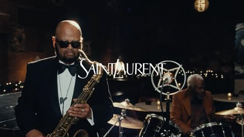 Two jazz musicians performing live on stage, one playing saxophone in the foreground and another one seated behind a drum kit.