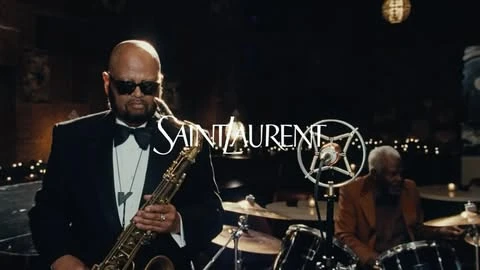 Two jazz musicians performing live on stage, one playing saxophone in the foreground and another one seated behind a drum kit.