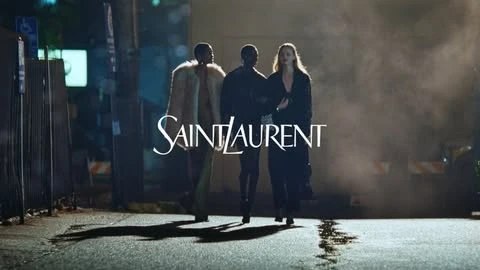 Three models walk down a dimly lit street in a Saint Laurent advertisement, showcasing fashion and urban style in a nighttime setting.