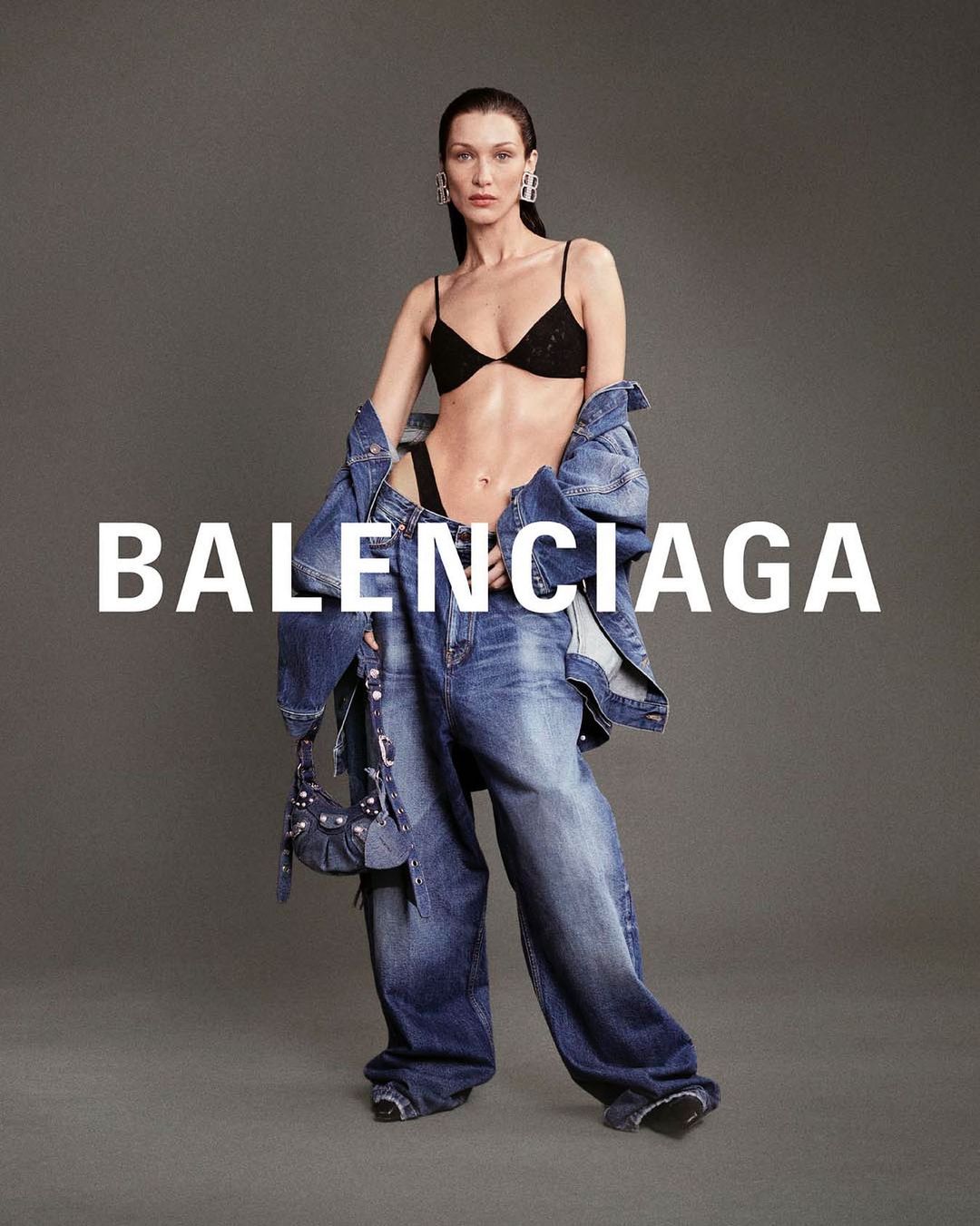 Bella Hadid poses in a Balenciaga ad, wearing a black bra and jeans. She holds a denim bag and stands against a grey background.