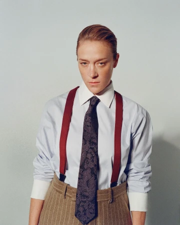 Portrait of a woman with slicked-back hair wearing a blue dress shirt, patterned tie, red suspenders, and brown pinstripe trousers.