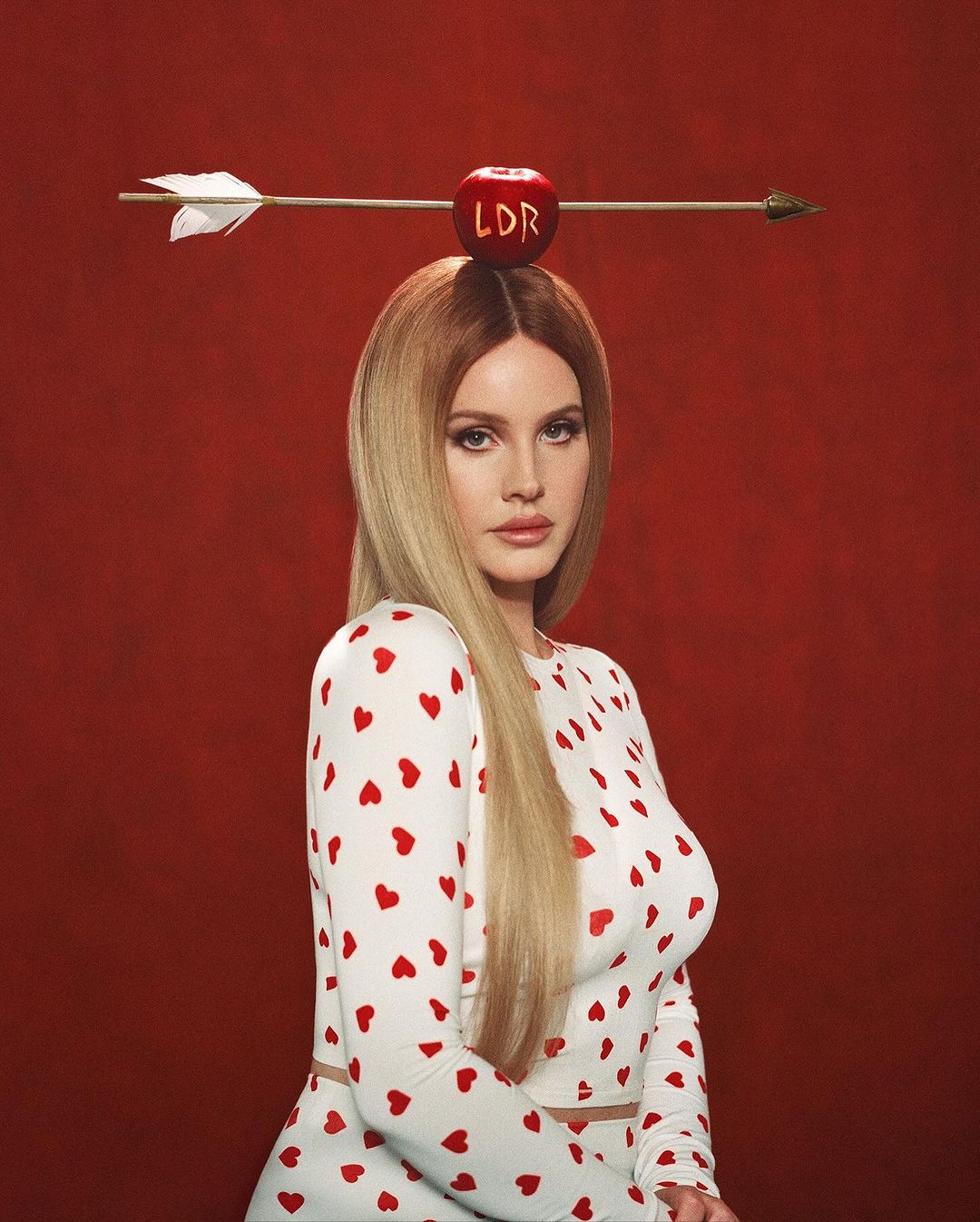 Portrait of a blonde woman with an apple pierced by an arrow on her head, wearing a white heart-print dress against a red backdrop.
