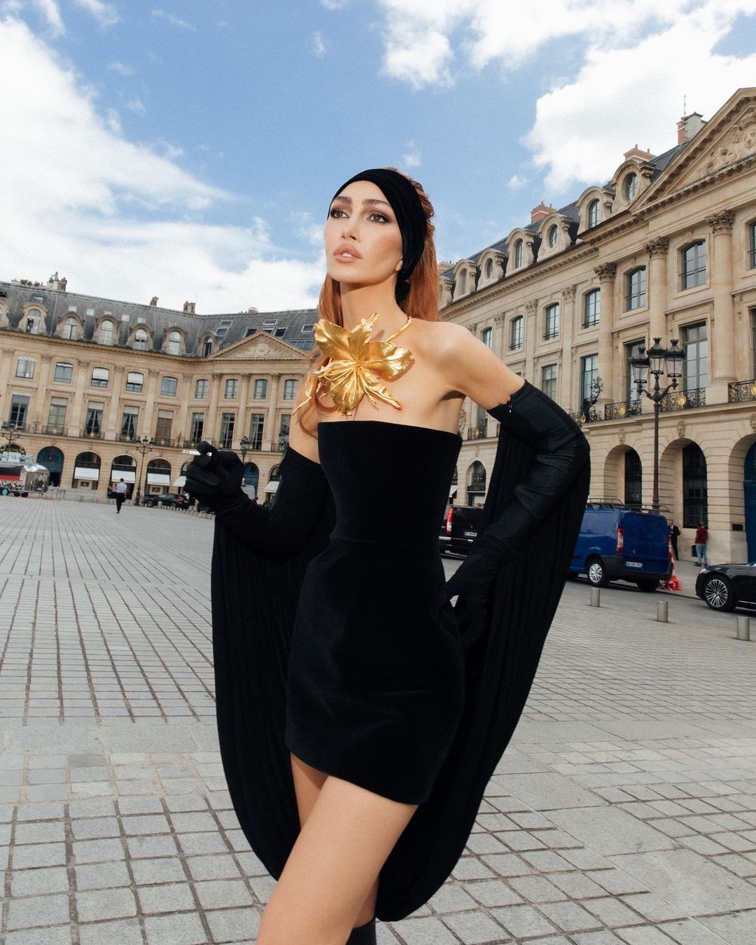 A woman in a black strapless dress and long gloves stands in a plaza with historic buildings under a partly cloudy sky.
