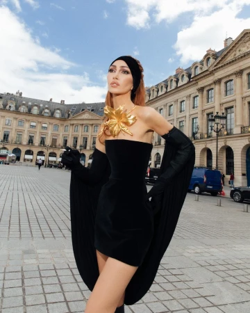 A woman in a black strapless dress and long gloves stands in a plaza with historic buildings under a partly cloudy sky.