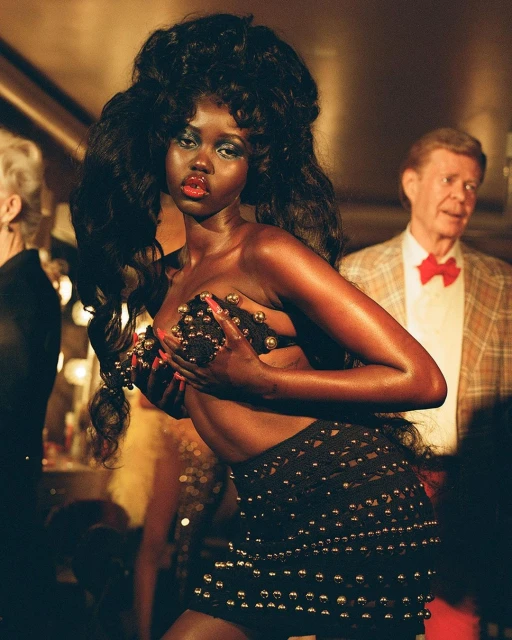 Striking dark-skinned woman poses in avant-garde fashion, adorned with textured hair and jeweled details, under warm, dramatic lighting.