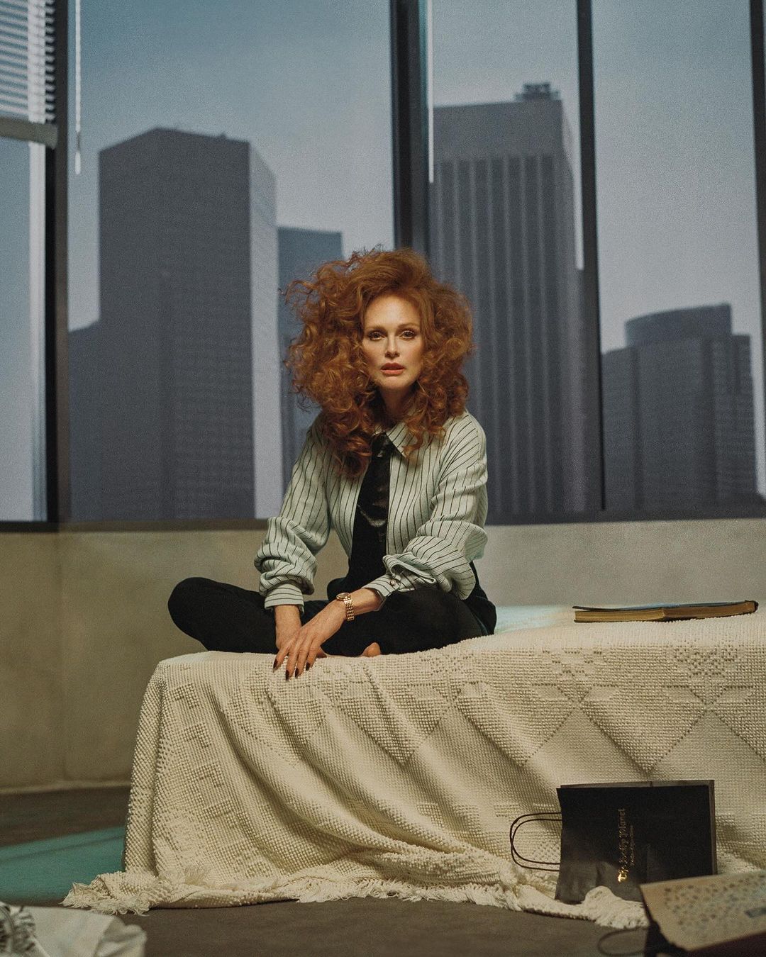 A redhead woman sits on a bed in front of a large window with skyscrapers. She wears a striped shirt and black pants.