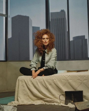 A redhead woman sits on a bed in front of a large window with skyscrapers. She wears a striped shirt and black pants.
