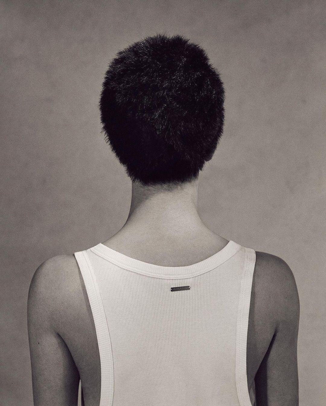 Back view of a person with short, dark hair wearing a white ribbed tank top against a neutral grey backdrop in a studio shot.